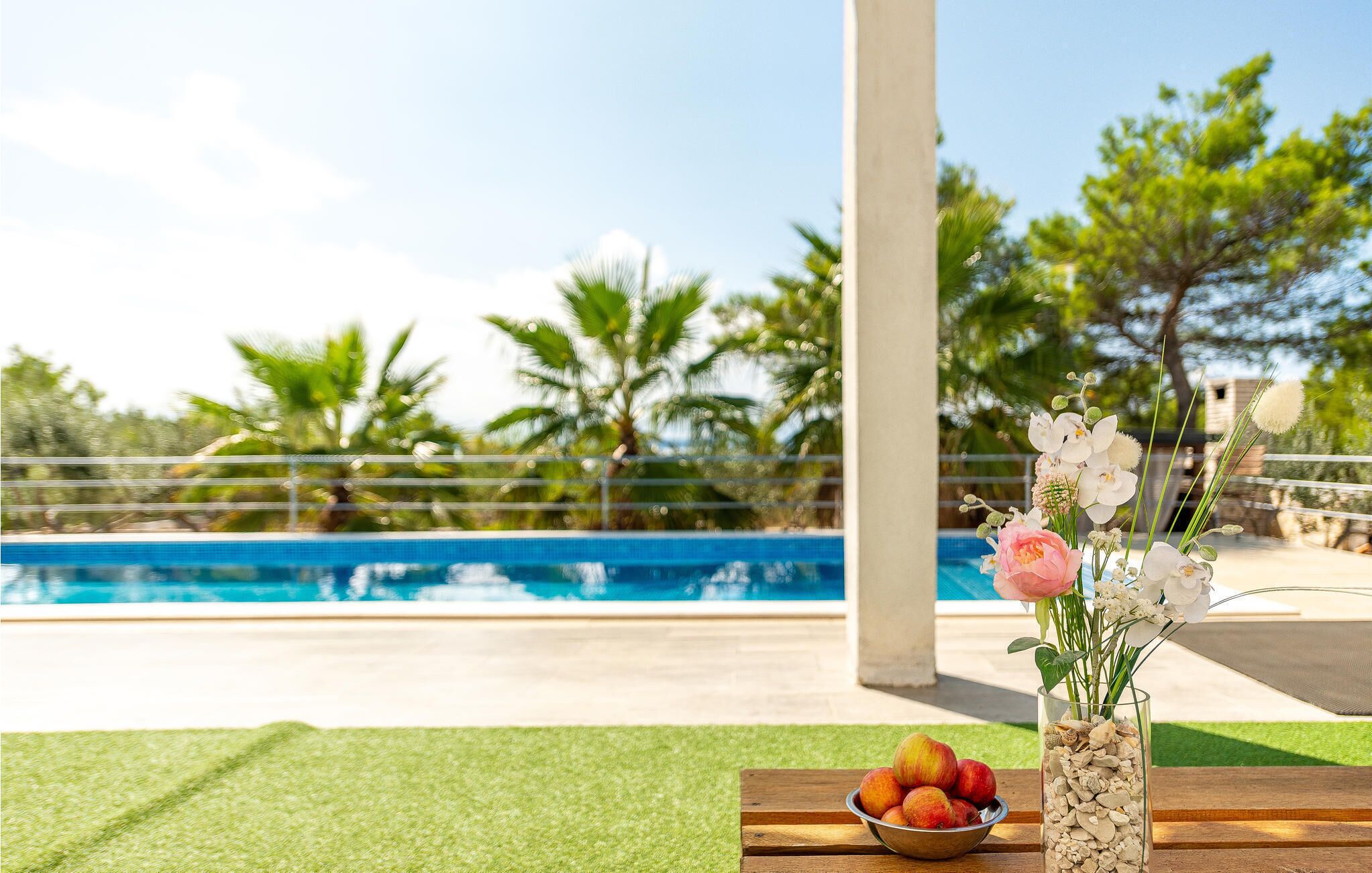 A poolside setting with a clear view of the pool, a wooden table with a bowl of fruit, and a vase with flowers.