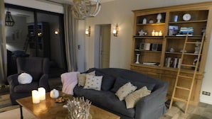Ein Wohnzimmer mit einem dunkelgrauen Sofa, einem Couchtisch aus Holz mit Kerzen, einem Sessel und einer Holzbibliothek mit verschiedenen Gegenständen.