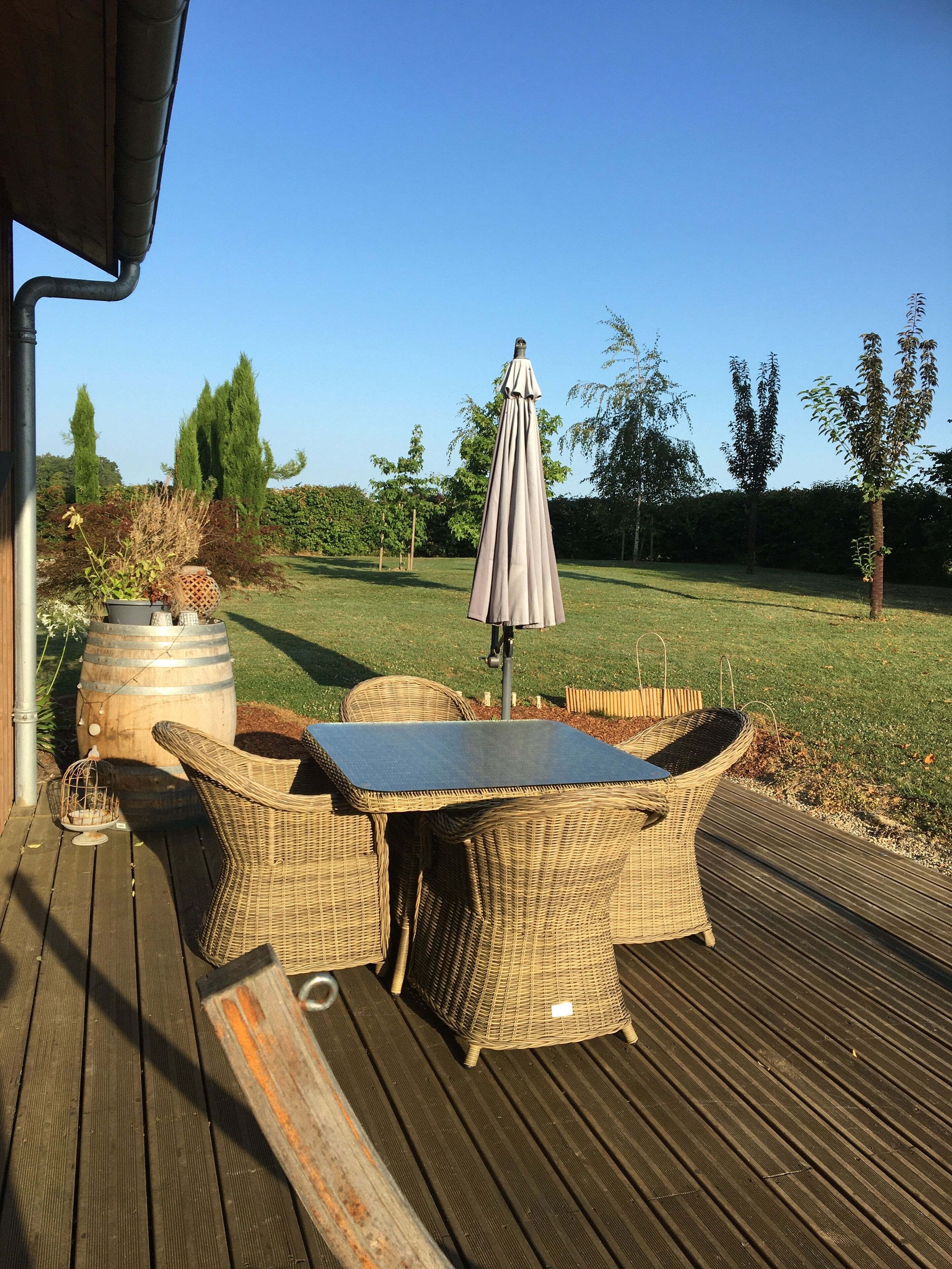 Una terrazza esterna con mobili in vimini e un tavolo, circondata da un patio in legno e una zona verde alberata.