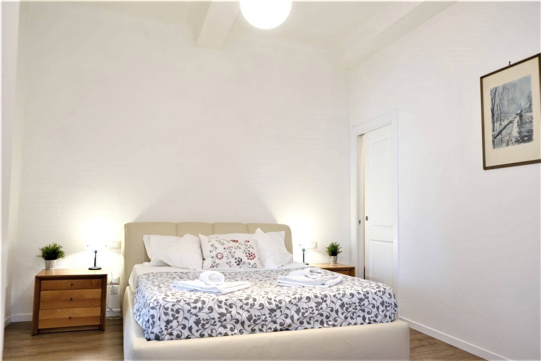Een slaapkamer met een bed, twee nachtkastjes, een nachtkastje, een lijst aan de muur en een deur.