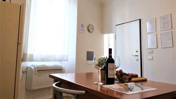 Ett litet, väl upplyst rum med ett träbord, en vinflaska och glas. Det finns ett fönster med tunna gardiner, en väggklocka och flera inramade dokument på väggen.