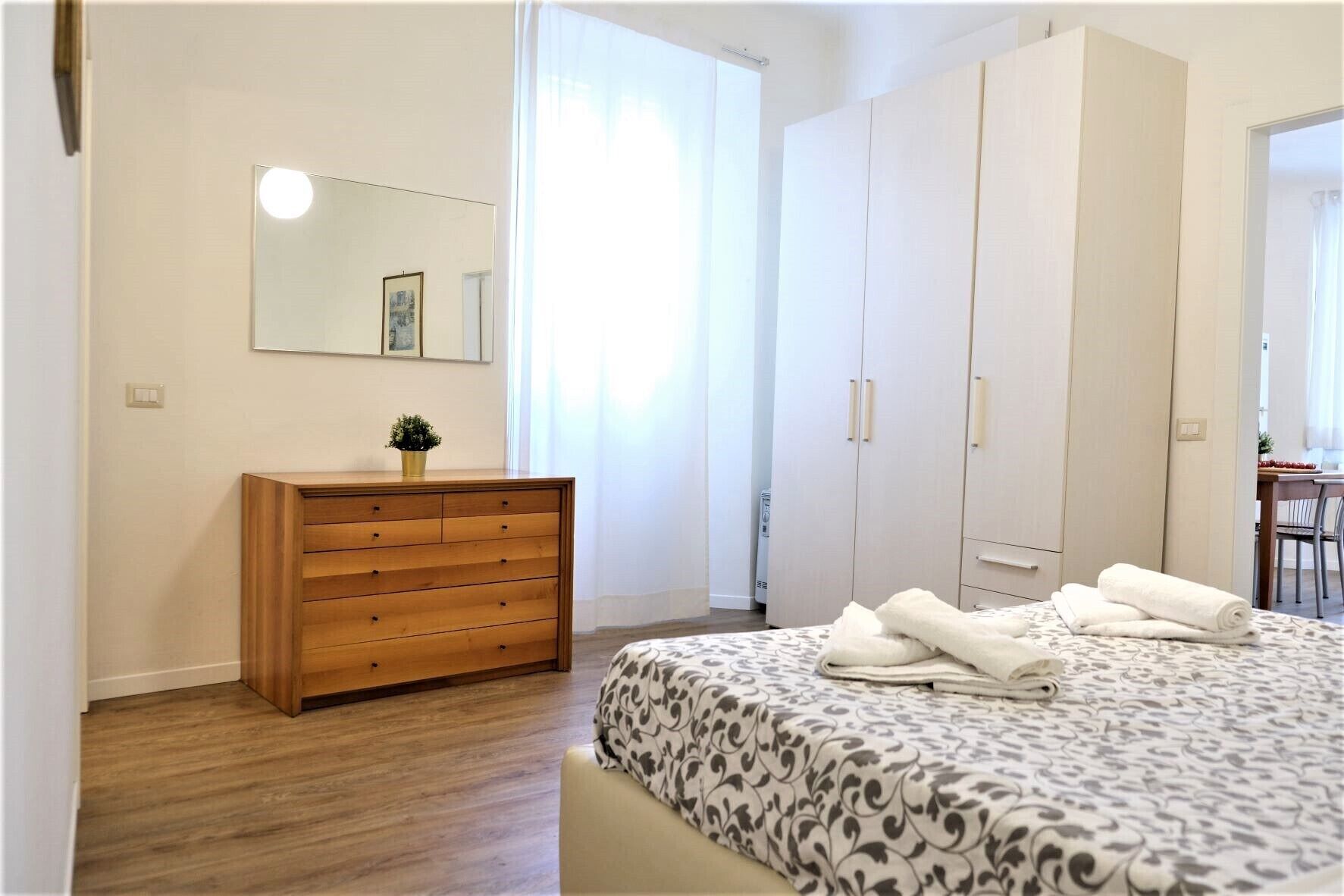 A bedroom with a wooden dresser, a bed with patterned bedding, a large wardrobe, and a mirror on the wall.
