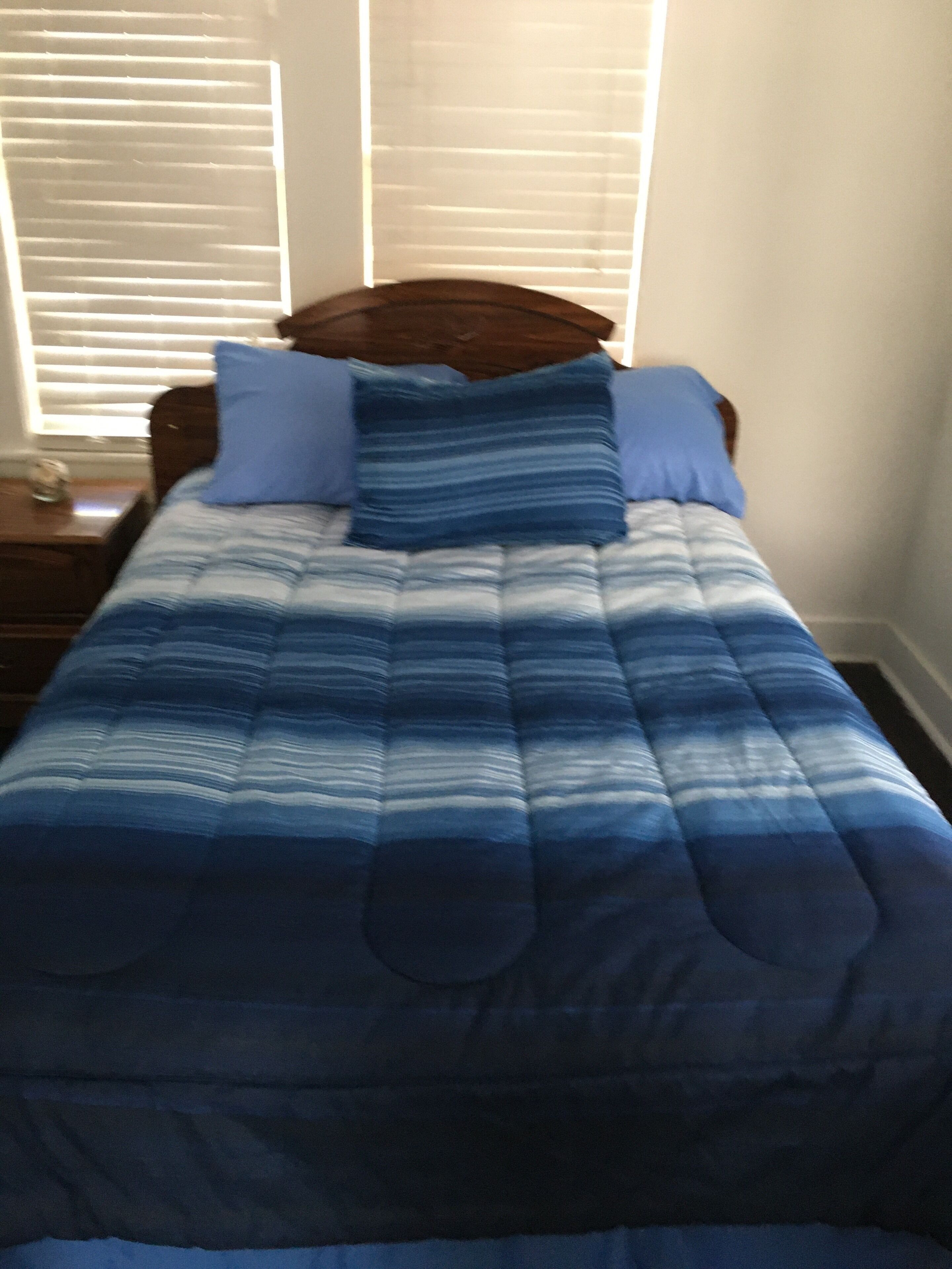 Un letto con copriletto azzurro a righe, due cuscini, una testiera in legno e un comodino con una lampada.