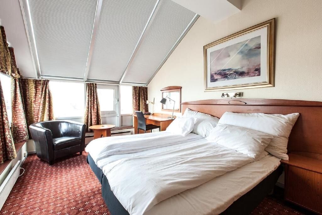 A hotel room with a large bed, a desk, a chair, and a window with curtains.