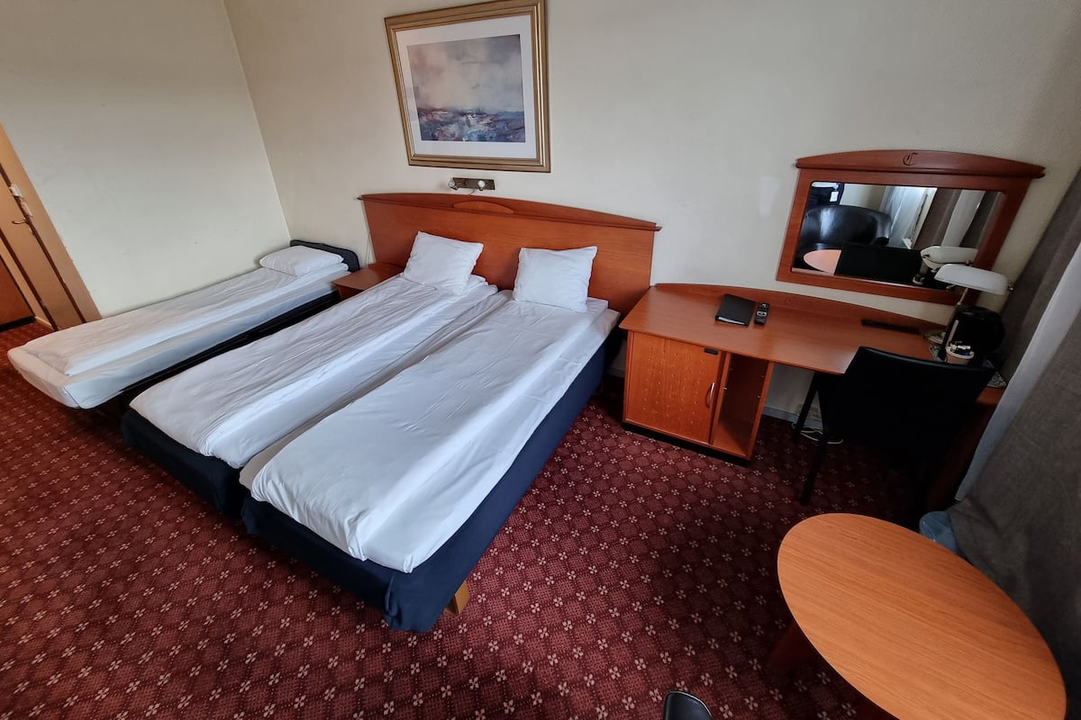 A hotel room with two single beds, a desk with a chair, a small cabinet, a bedside table, a mirror, and a painting on the wall.