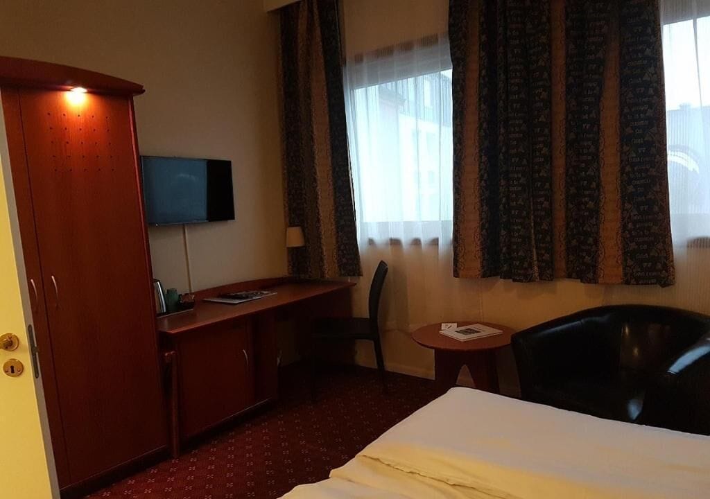 Quarto de hotel com cama, escrivaninha, cadeira, televisão e janela com cortinas.