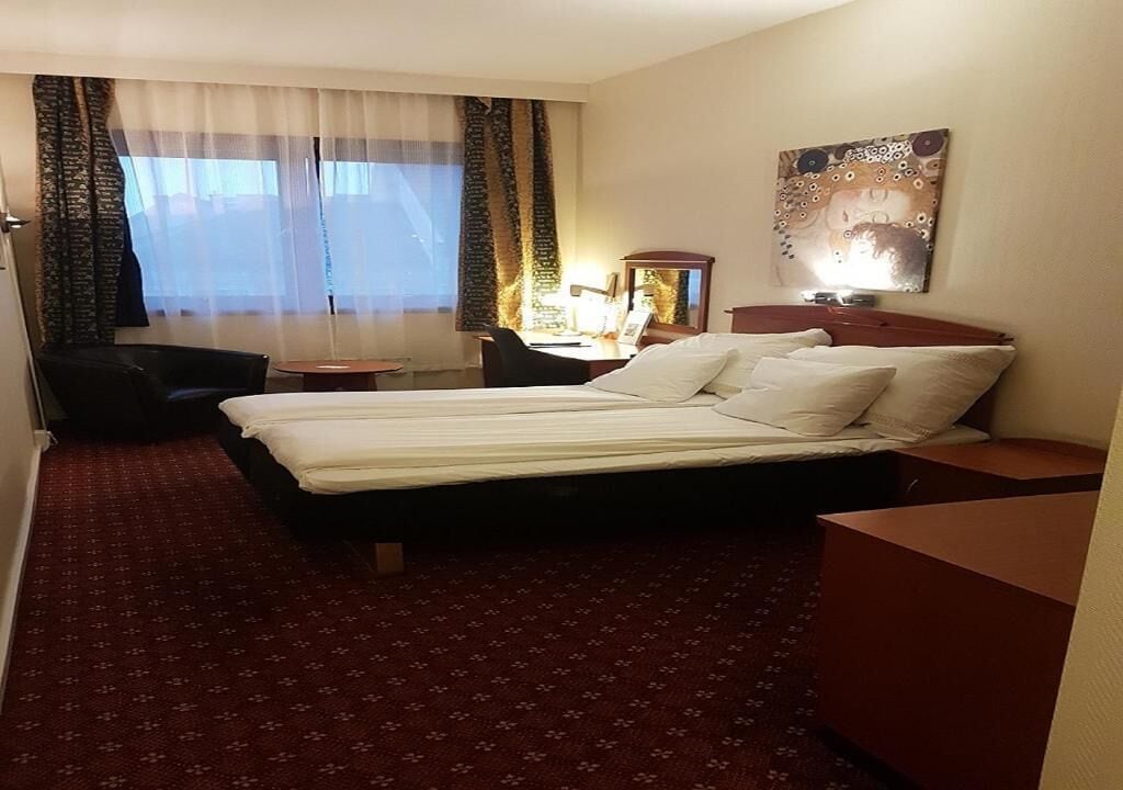 Habitación de hotel con cama, escritorio con silla, una mesita y un cuadro en la pared.