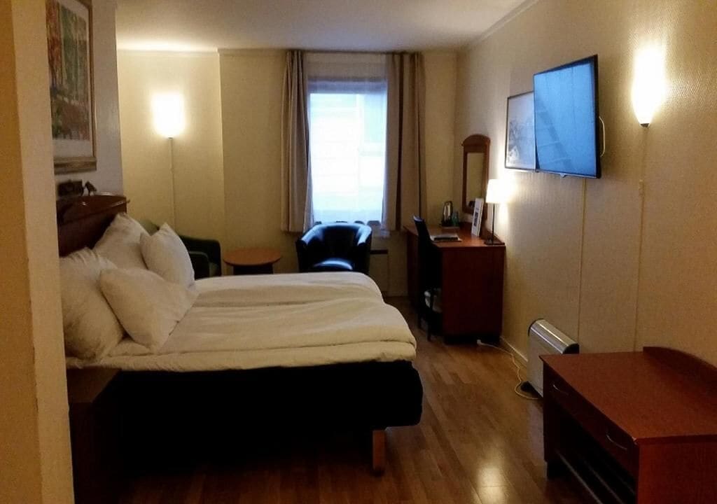 A hotel room with a double bed, a desk with a chair, a TV mounted on the wall, and a window with curtains.