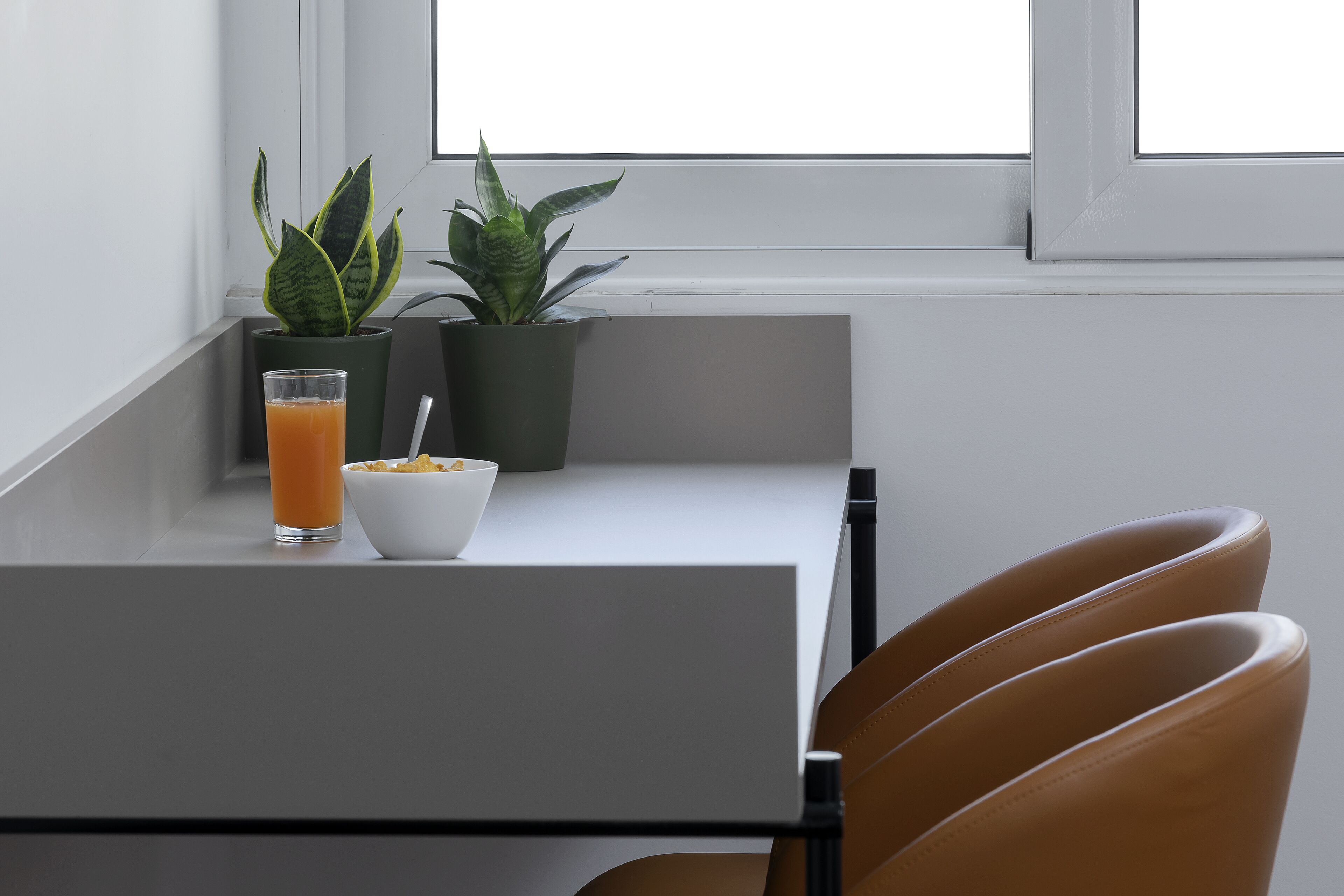 Un comedor con una mesa, dos plantas en macetas, un vaso de zumo de naranja y un bol de cereales.