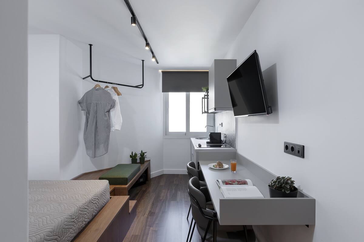 A compact, modern bedroom with a bed, bedside table, and a desk area with a chair, a flat-screen TV, and a small potted plant.