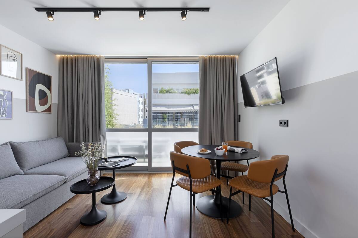 A modern living room with a grey sofa, a round dining table with chairs, a flat-screen TV mounted on the wall, and large windows with curtains.