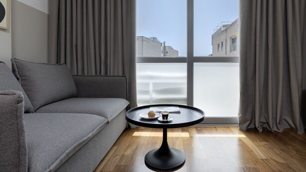 Un saló modern amb un sofà gris, una taula rodona negra amb una tassa i un pastís, i una finestra gran amb cortines.