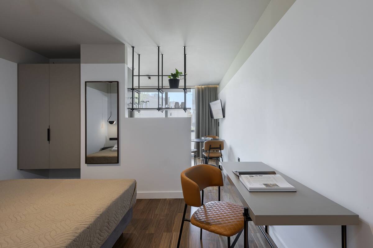 A modern bedroom with a bed, a desk, and a chair. There is a window with curtains, a potted plant, and a mirror on the wall.