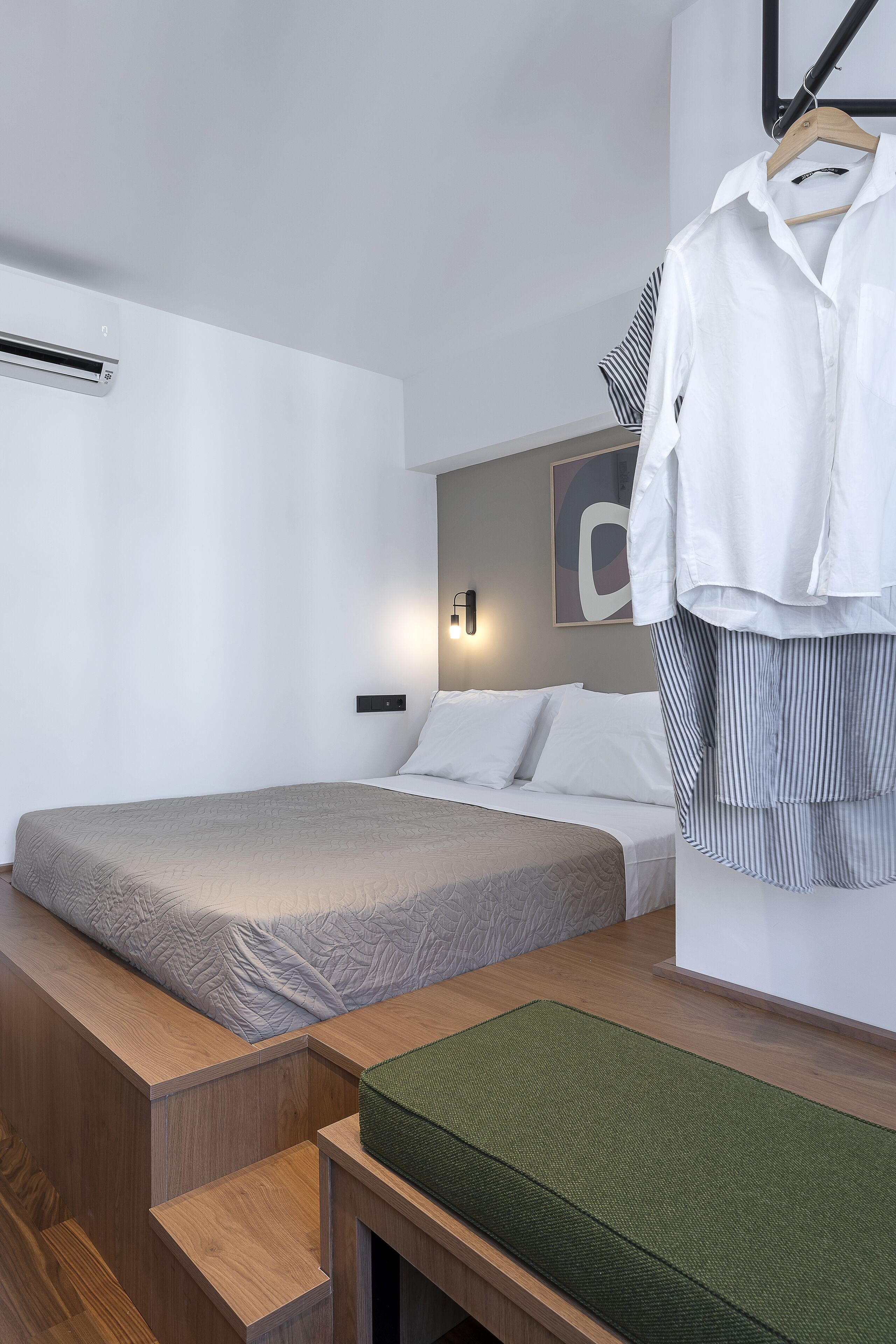 Une chambre d’hôtel moderne avec un lit, un banc en bois, un tapis vert et une chemise blanche suspendue à un porte-vêtements.