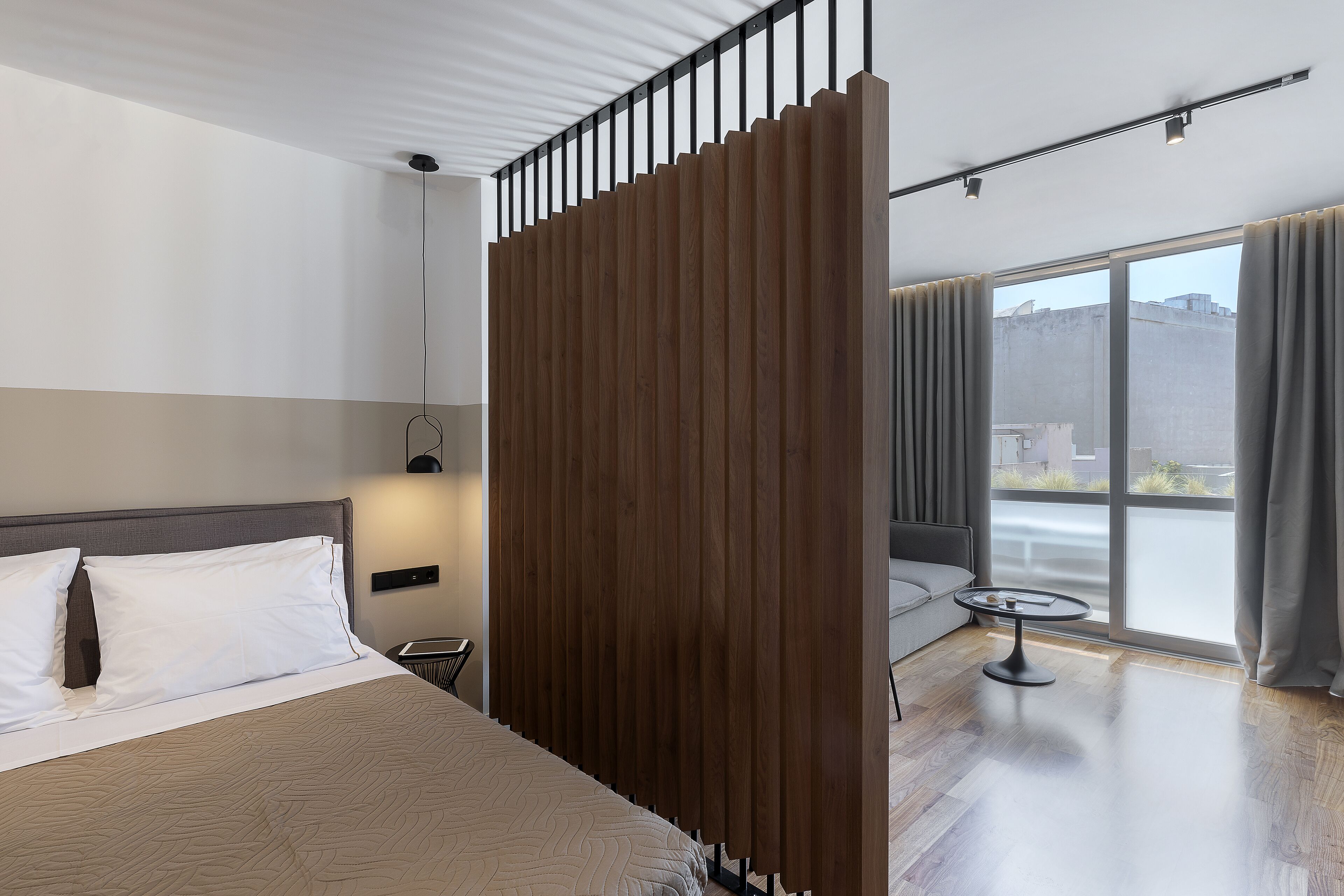 Una camera d'albergo moderna con un letto grande, un pannello in legno, un tavolino e vista sulla città attraverso ampie finestre.