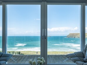 Een kamer met een grote glazen deur die uitkomt op een balkon met uitzicht op de oceaan en een rotsachtige kustlijn.