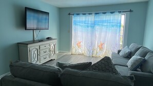 A living room with a flat-screen TV, a white cabinet, a grey sofa, and a window with a starfish curtain. - Just bring your bathing suit ! Steps to the beach ! (Myrtle Beach)