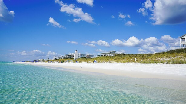 Uma praia com águas turquesa cristalinas, areia branca e alguns edifícios ao fundo.