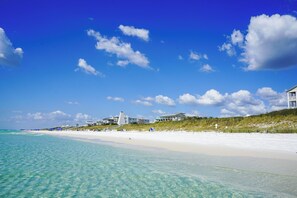 Une plage avec une eau turquoise et limpide, du sable blanc et quelques bâtiments en arrière-plan.
