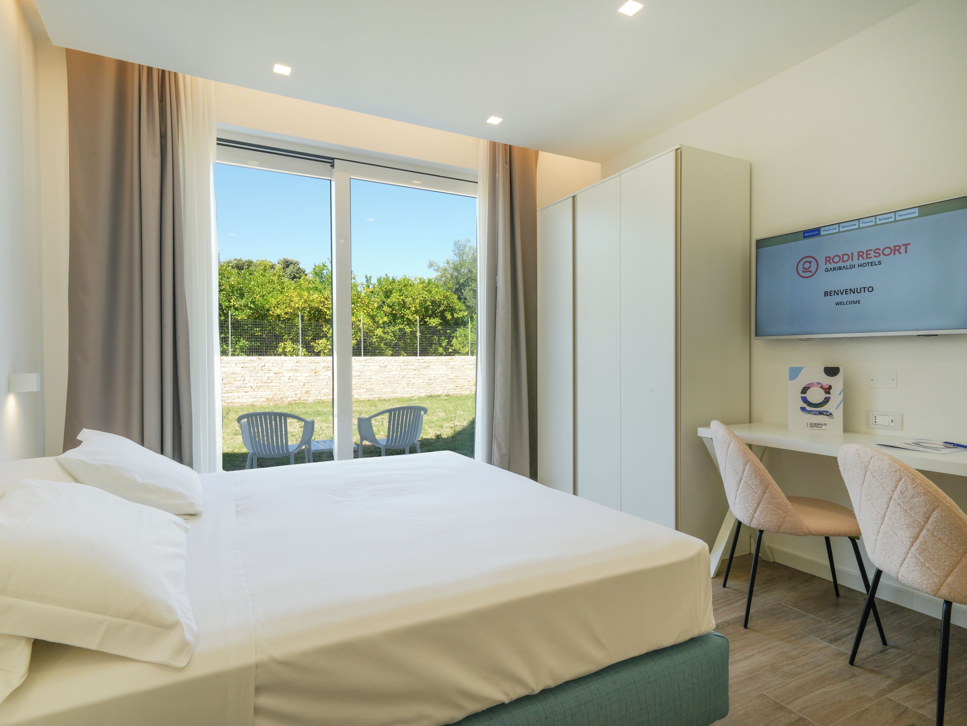 Et hotelværelse med seng, skrivebord, stol og et fjernsyn, der viser hotellets navn.