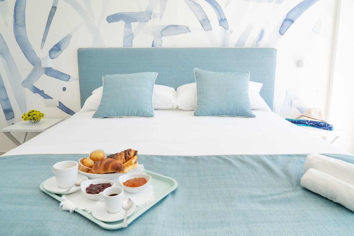 A neatly made bed with a blue headboard, a tray with croissants, coffee, and jam, and a small vase with yellow flowers on a white nightstand.