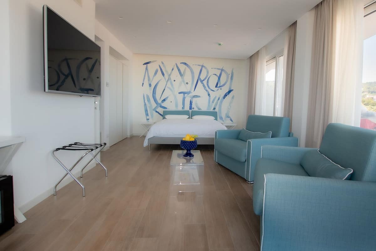 A modern hotel room with a bed, two blue armchairs, a TV, and a large window with a view.