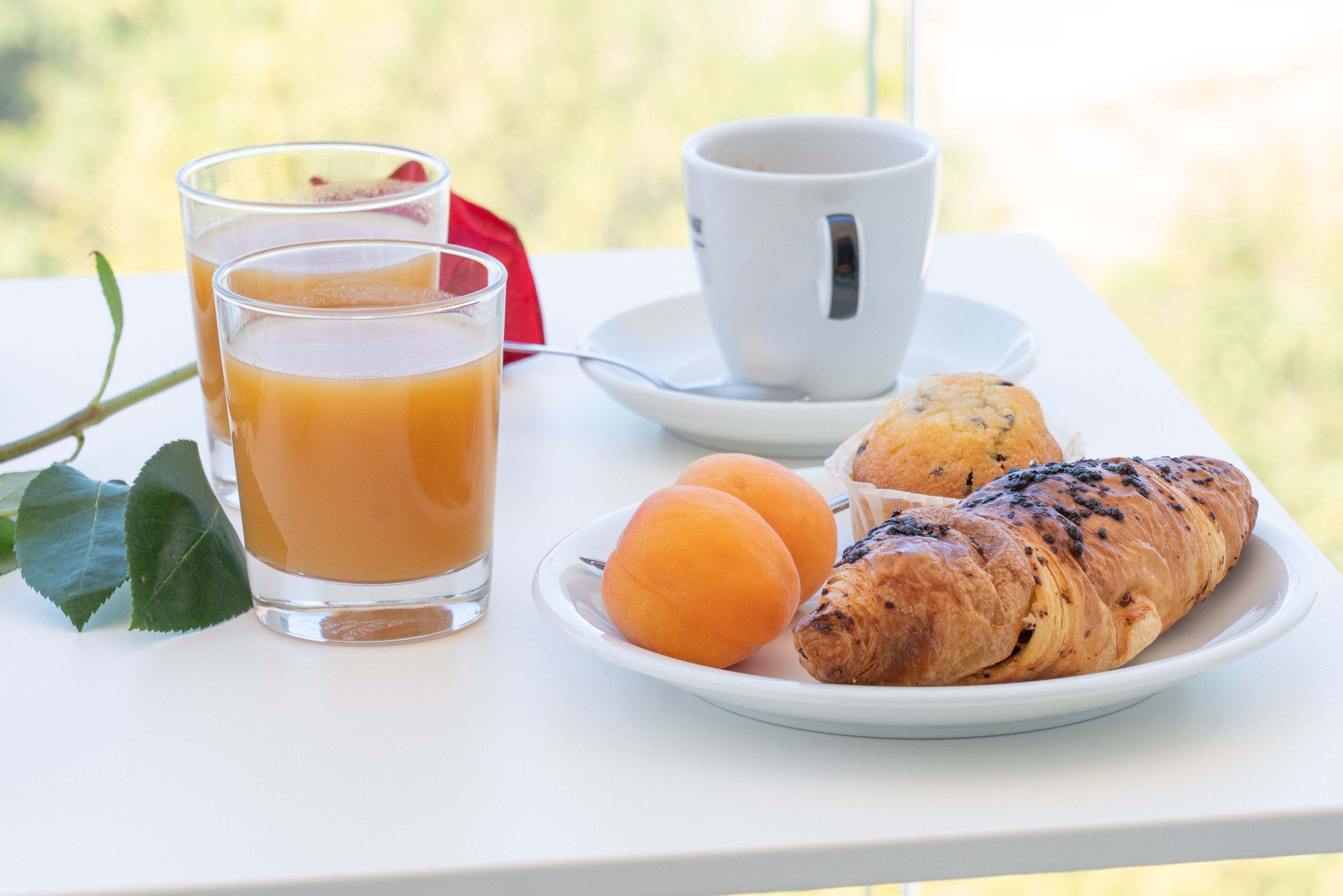 A breakfast setting with a glass of orange juice, a cup of coffee, a croissant, and two apricots on a white table.