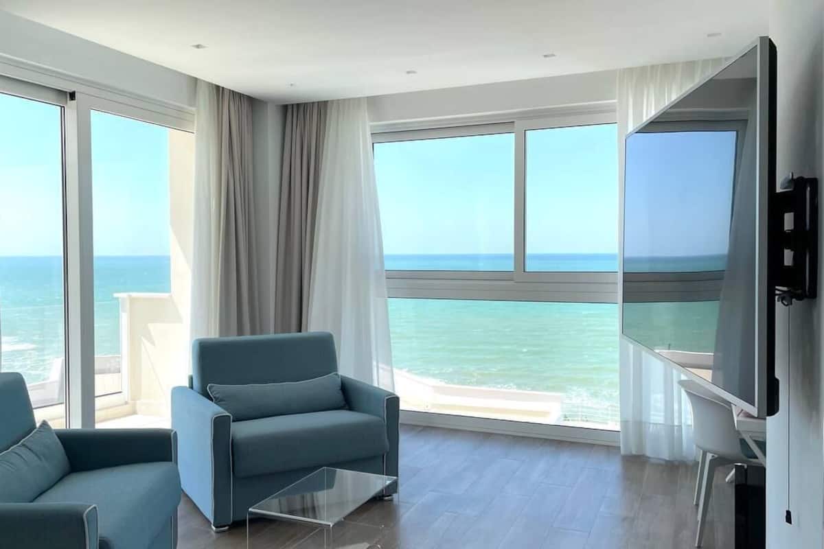 A modern hotel room with a large balcony, a sofa, a TV, and a glass coffee table.