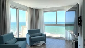 A modern hotel room with a large balcony, a sofa, a TV, and a glass coffee table.