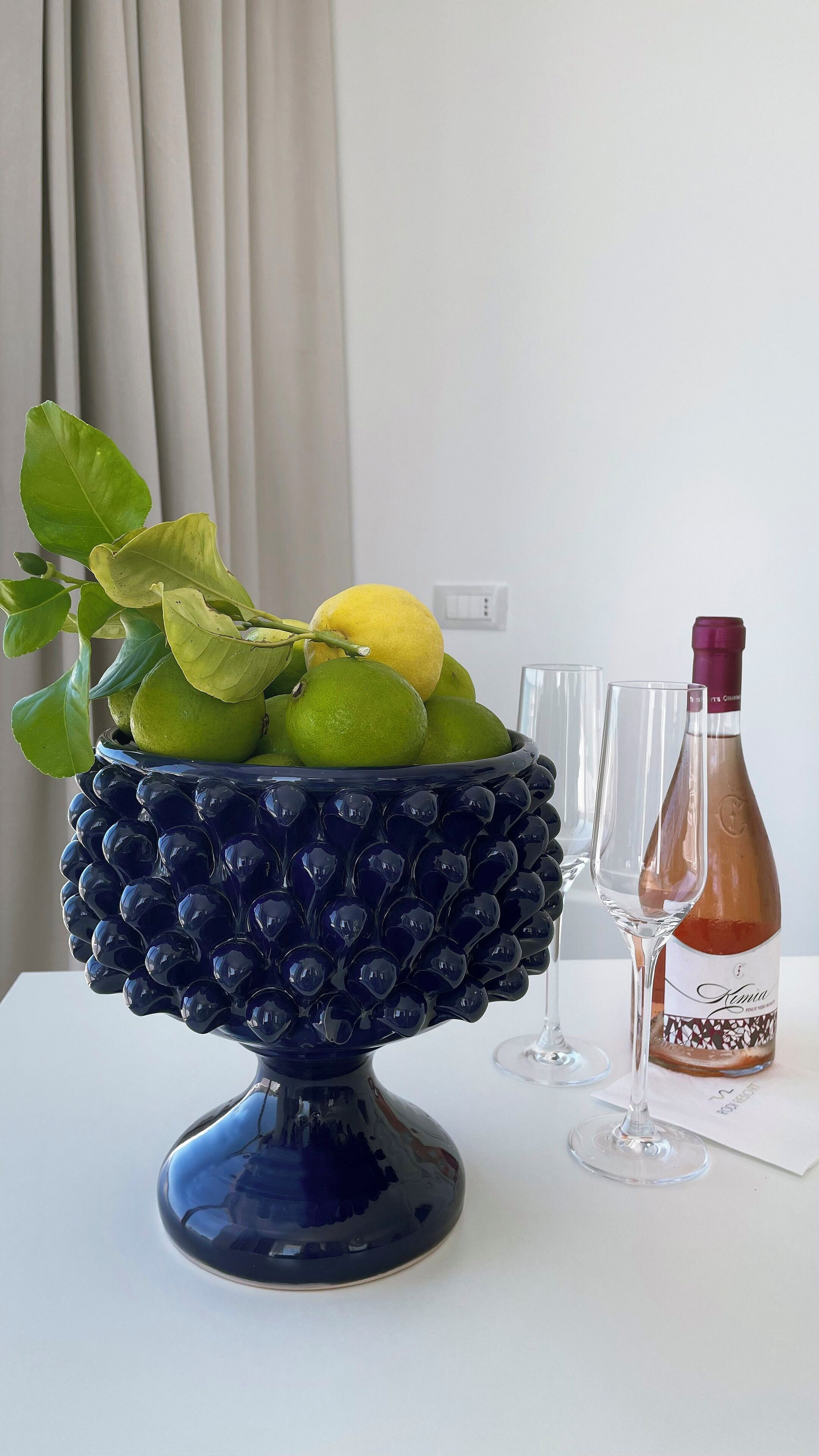 A decorative blue bowl filled with green and yellow citrus fruits, a bottle of red wine, and two empty wine glasses on a white table.