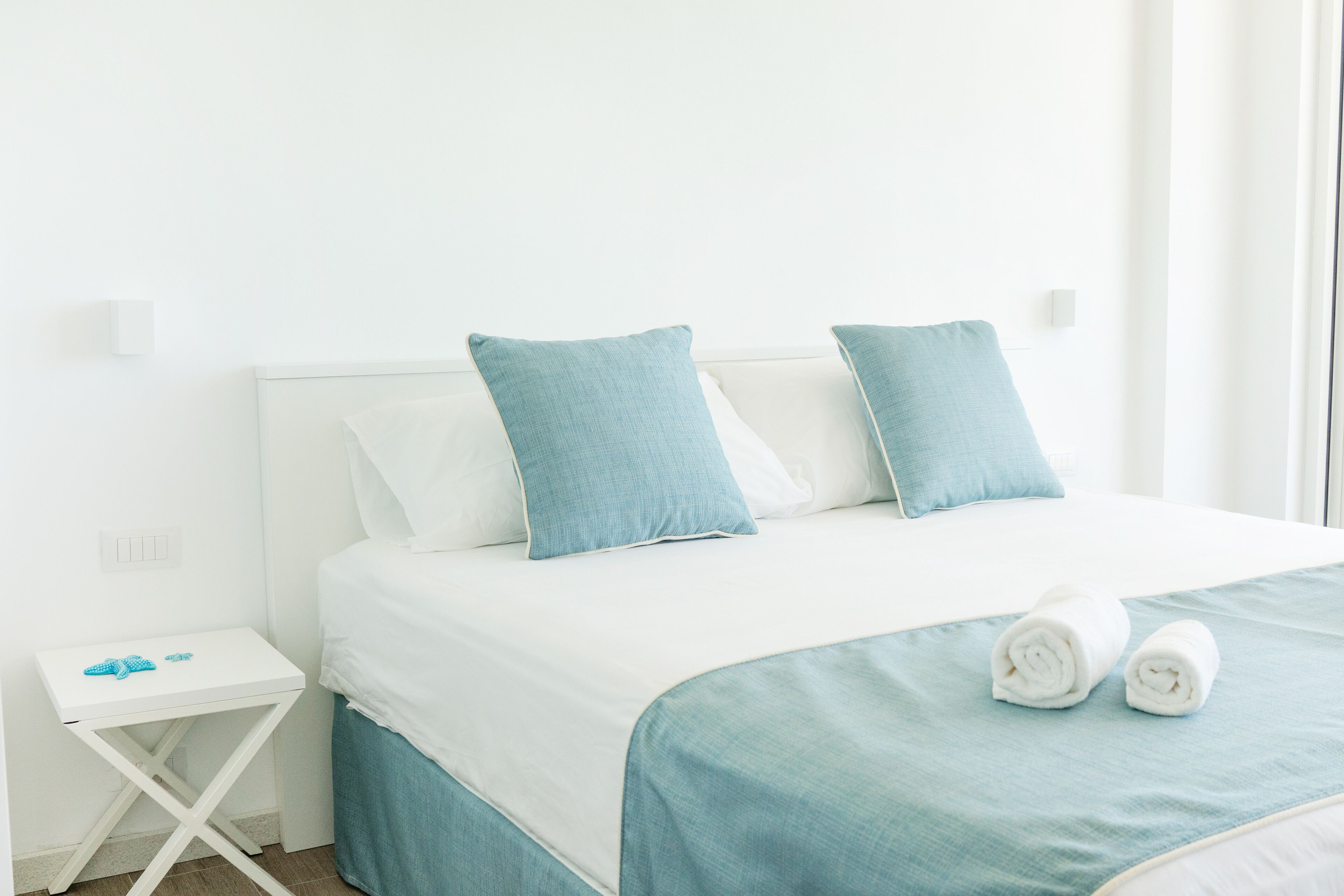 Un lit bien fait, avec une literie blanche et bleue, une petite table de chevet sur laquelle se trouve un objet bleu, et une commode blanche.