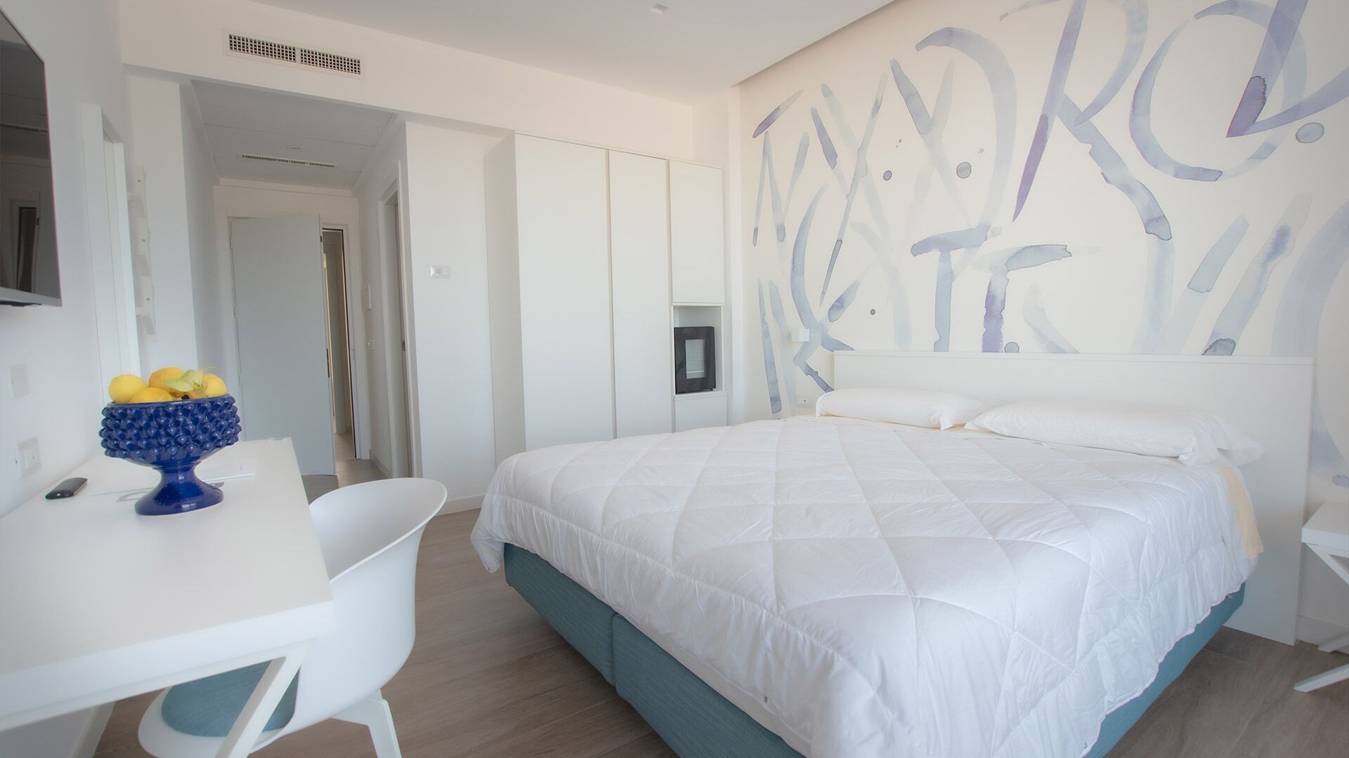 A modern bedroom with a large bed, a desk, and a chair. There is a wall art piece above the bed.