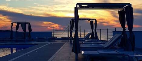 A rooftop terrace with sun loungers and a pool, overlooking the sunset.