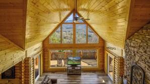 A log cabin interior with a wooden ceiling, a television on a wooden stand, a stone wall, and a balcony with a view of the mountains. - Priceless Views, Game Room, Hot Tub, Firepit & Newly Remodeled! Free Tickets! (Gatlinburg)