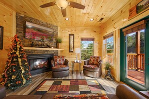 A cozy cabin interior with a fireplace, two leather armchairs, a Christmas tree, and a large window offering a view of the outdoors. - Free Attractions | Indoor Heated Pool, Game Room, Fireplace, 2 Miles to Strip (Sevierville)
