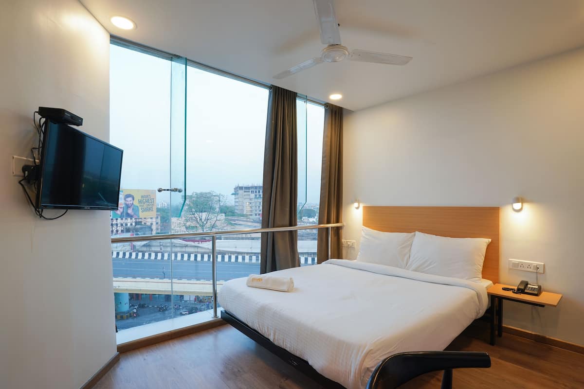 A hotel room with a bed, a TV mounted on the wall, a bedside table with a phone, and a view of the city through large windows.
