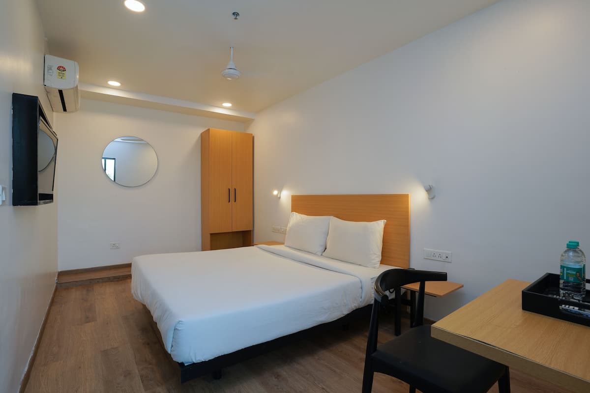 A hotel room with a bed, a wooden headboard, a round mirror, a small table with a chair, and a water bottle on the table.