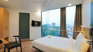 Quarto de hotel moderno com uma cama grande, uma escrivaninha, uma cadeira, uma televisão presa à parede e vista da cidade através de amplas janelas.