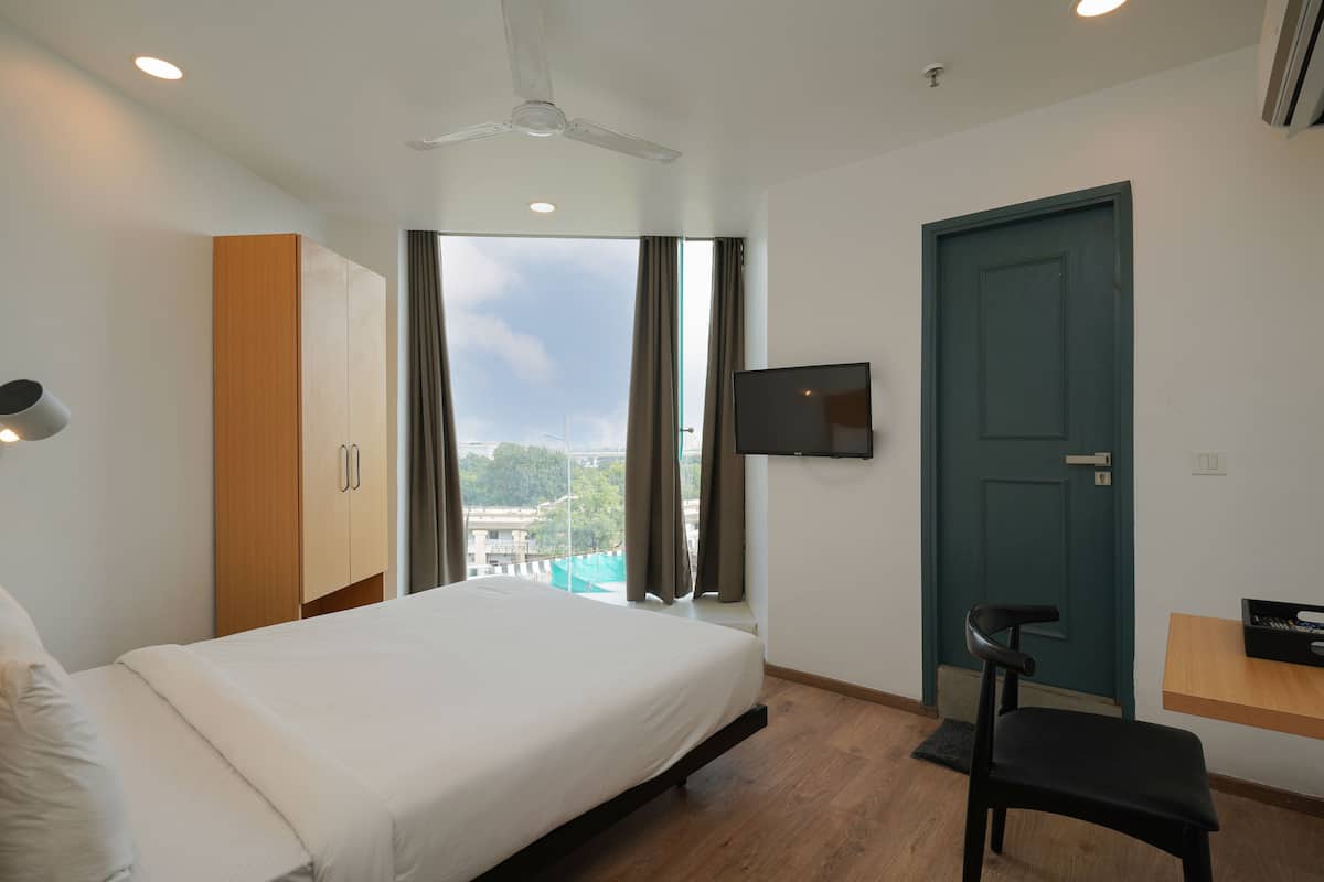 A modern hotel room with a bed, wardrobe, TV, desk, chair, and a view of the outdoors.