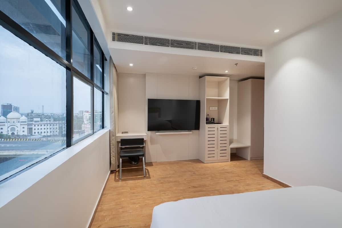 A modern hotel room with a large window, a flat-screen TV, a bed, and a wardrobe.