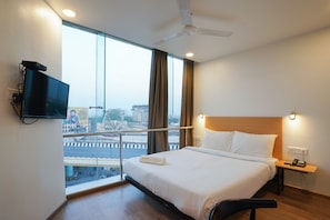 A hotel room with a bed, a television, a nightstand, a telephone, and a view of the city through large windows. - Uptown Hotel (Nagpur)