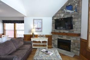 A living room with a stone fireplace, a flat-screen TV mounted above it, a brown leather sofa, and a patterned ottoman.