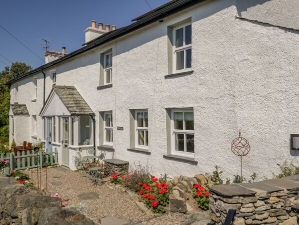 A white two-story house with a stone foundation, multiple windows, and a small garden with red flowers. - 1 Sunny Point Cottages (Kendal)
