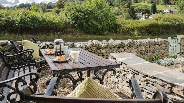 A rustic outdoor seating area with a stone wall, wooden table, and metal chairs. - 1 Sunny Point Cottages (Kendal)