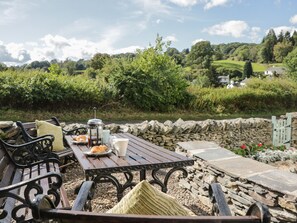 A rustic outdoor seating area with a stone wall, wooden table, and metal chairs. - 1 Sunny Point Cottages (Kendal)