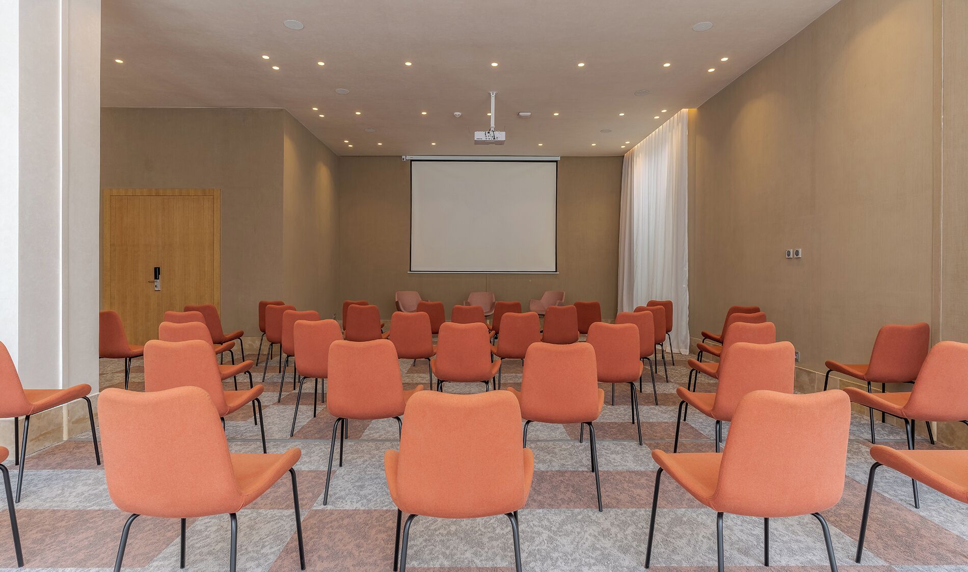 Uma sala de conferências com fileiras de cadeiras laranja, uma tela de projeção grande e uma porta de madeira.