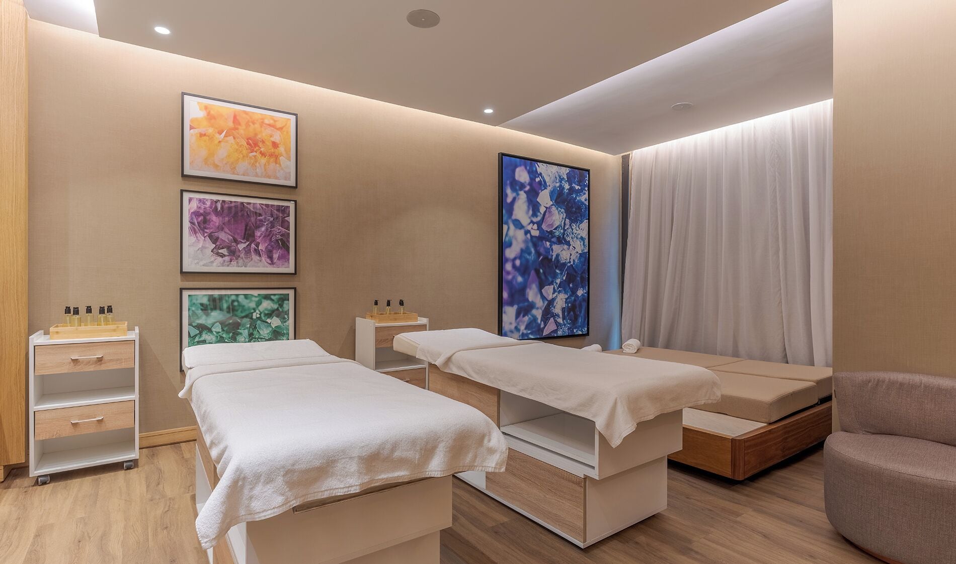 Une salle de spa équipée de deux tables de massage, d’un meuble en bois contenant des bouteilles, et de trois tableaux abstraits encadrés accrochés au mur.