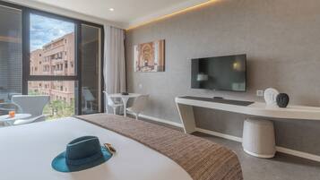 A modern hotel room with a large bed, a TV, a desk, and a view of the city through large windows.