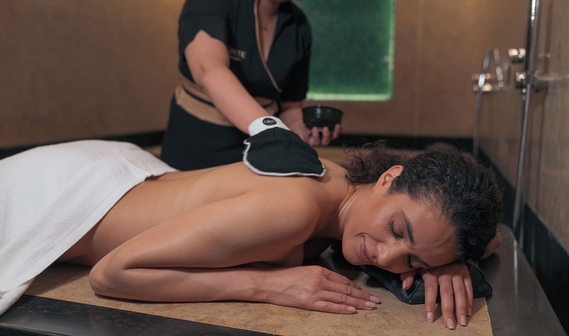 A person receiving a back massage in a room with a wooden floor and a green towel hanging on the wall.