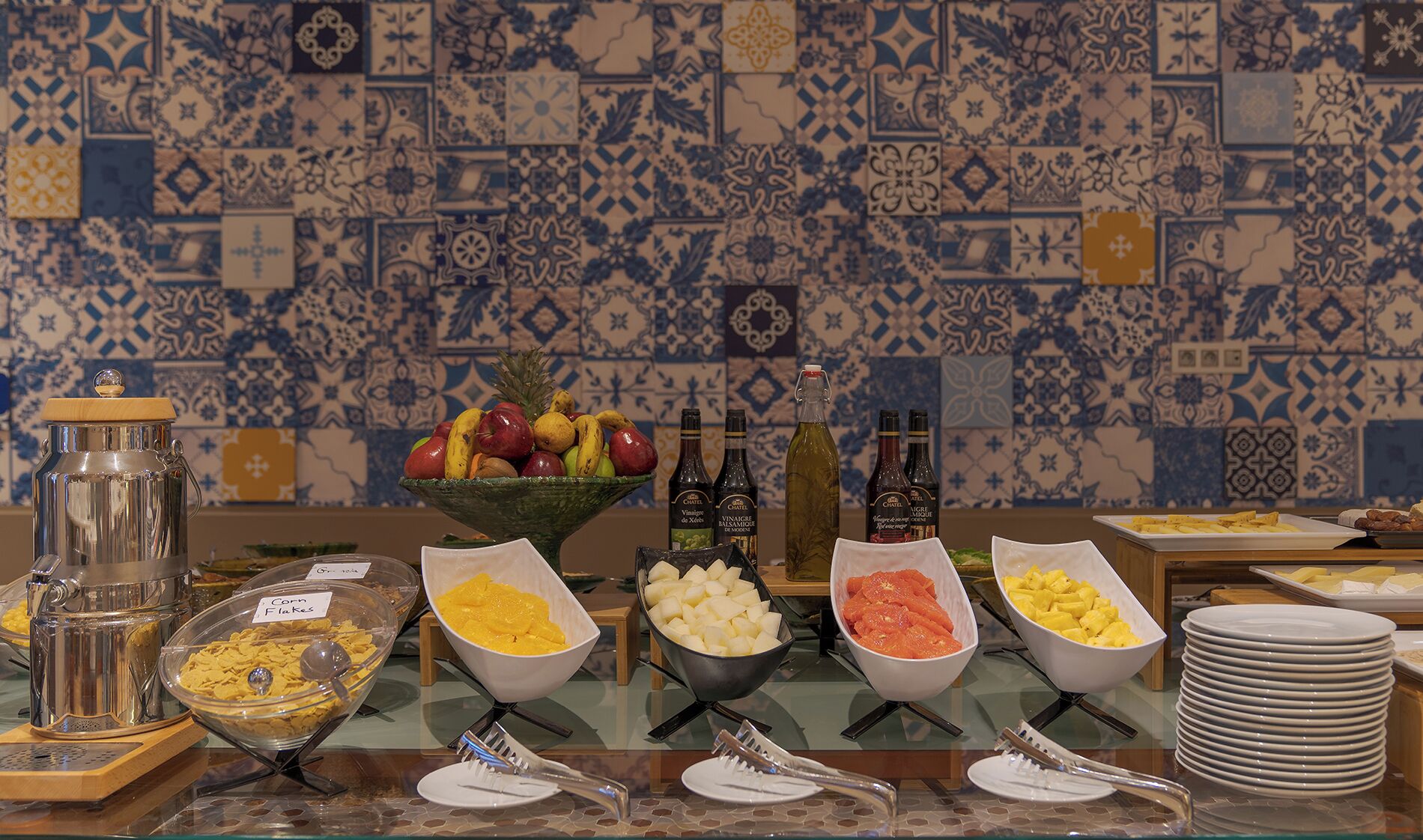 Un buffet proposant une variété de plats et de boissons, notamment des fruits, des fromages et des condiments, le tout disposé devant un mur carrelé.